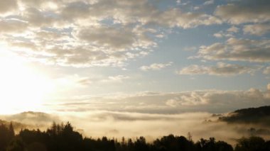 Altın gündoğumunda bulutlar ve dağlar. Sabah sisi, çam ağaçlarının üzerinde, güneş ışığıyla. Beyaz kabarık bulutların zaman akışı sabah gökyüzünde hareket ederken arkaplanda siluet dağları var.