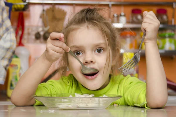 Una niña de cuatro años come con un tenedor y una cuchara sentada en la mesa de la cocina —  Fotos de Stock