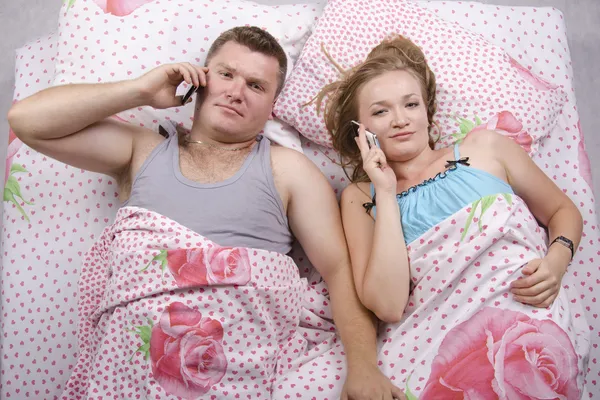 Mladý pár mluvit do telefonu, ležící v posteli — Stock fotografie