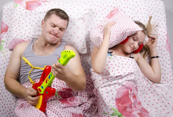 Pár v posteli manžel hraje na kytaru, manželka pokrytá polštáři — Stock fotografie
