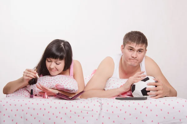 Girl paints her nails in bed, man emotionally watching soccer — Stock Photo, Image