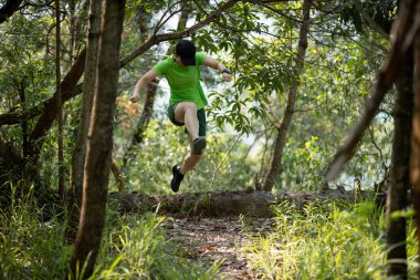 Woman trail runner running and jumping over a fallen tree in tropical forest mountain 