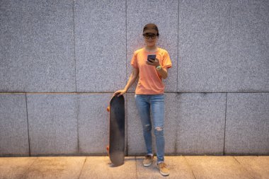 Female skateboarder using smartphone leaning on wall in city