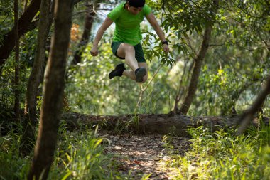 Woman trail runner running and jumping over a fallen tree trunk in tropical forest mountain