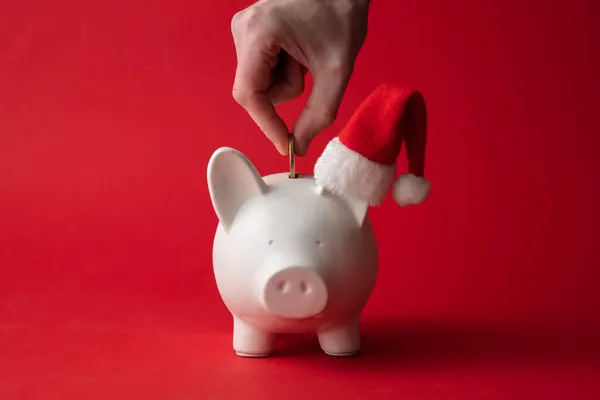 Χέρι βάζοντας χρήματα σε ένα κουμπαρά φορώντας ένα καπέλο Σάντα. Χριστουγεννιάτικη αποταμίευση — Φωτογραφία Αρχείου