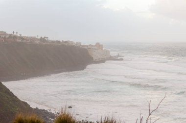 Kayalıklardan gelen dalgalı deniz manzarası, kıyıya vuran bir sürü beyaz köpüklü büyük dalgalar, fırtına bulutları ve resimli balıkçı köyü. Bajamar, Tenerife, Kanarya Adaları. İspanya