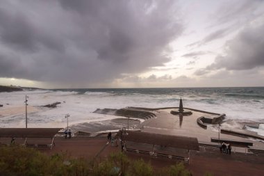Büyük dalgaların fırtınası dalgakırana karşı kırılıyor ve Bajamar geziniyor gri fırtına bulutları ve arka planda bir deniz feneri ile.