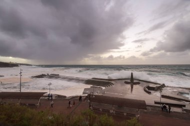 Büyük dalgaların fırtınası dalgakırana karşı kırılıyor ve Bajamar geziniyor gri fırtına bulutları ve arka planda bir deniz feneri ile.