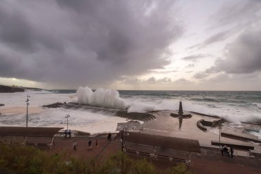 Büyük dalgaların fırtınası dalgakırana karşı kırılıyor ve Bajamar geziniyor gri fırtına bulutları ve arka planda bir deniz feneri ile.
