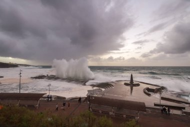 Büyük dalgaların fırtınası dalgakırana karşı kırılıyor ve Bajamar geziniyor gri fırtına bulutları ve arka planda bir deniz feneri ile.