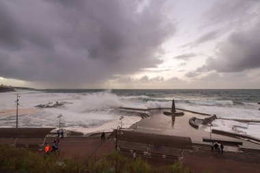 Büyük dalgaların fırtınası dalgakırana karşı kırılıyor ve Bajamar geziniyor gri fırtına bulutları ve arka planda bir deniz feneri ile.