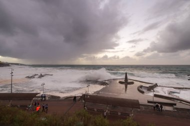 Büyük dalgaların fırtınası dalgakırana karşı kırılıyor ve Bajamar geziniyor gri fırtına bulutları ve arka planda bir deniz feneri ile.
