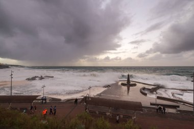 Büyük dalgaların fırtınası dalgakırana karşı kırılıyor ve Bajamar geziniyor gri fırtına bulutları ve arka planda bir deniz feneri ile.