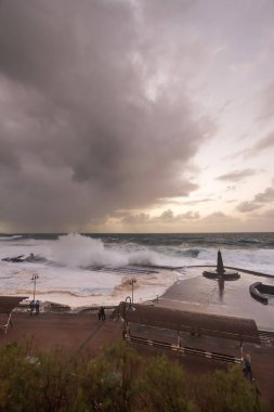 Büyük dalgaların fırtınası dalgakırana karşı kırılıyor ve Bajamar geziniyor gri fırtına bulutları ve arka planda bir deniz feneri ile.