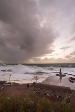 Büyük dalgaların fırtınası dalgakırana karşı kırılıyor ve Bajamar geziniyor gri fırtına bulutları ve arka planda bir deniz feneri ile.