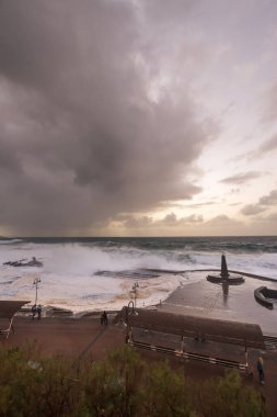Büyük dalgaların fırtınası dalgakırana karşı kırılıyor ve Bajamar geziniyor gri fırtına bulutları ve arka planda bir deniz feneri ile.