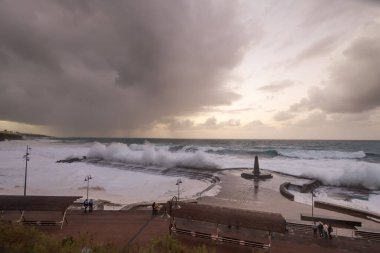 Büyük dalgaların fırtınası dalgakırana karşı kırılıyor ve Bajamar geziniyor gri fırtına bulutları ve arka planda bir deniz feneri ile.