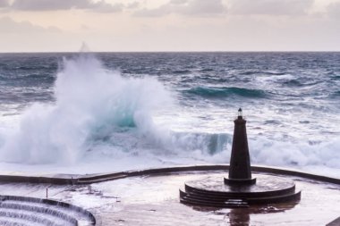 Büyük dalgaların fırtınası dalgakırana karşı kırılıyor ve Bajamar geziniyor gri fırtına bulutları ve arka planda bir deniz feneri ile.