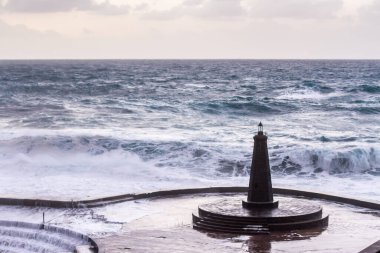 Büyük dalgaların fırtınası dalgakırana karşı kırılıyor ve Bajamar geziniyor gri fırtına bulutları ve arka planda bir deniz feneri ile.
