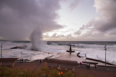 Büyük dalgaların fırtınası dalgakırana karşı kırılıyor ve Bajamar geziniyor gri fırtına bulutları ve arka planda bir deniz feneri ile.