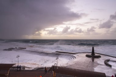 Büyük dalgaların fırtınası dalgakırana karşı kırılıyor ve Bajamar geziniyor gri fırtına bulutları ve arka planda bir deniz feneri ile.