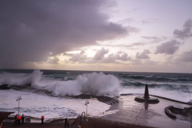 Büyük dalgaların fırtınası dalgakırana karşı kırılıyor ve Bajamar geziniyor gri fırtına bulutları ve arka planda bir deniz feneri ile.