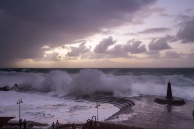 Büyük dalgaların fırtınası dalgakırana karşı kırılıyor ve Bajamar geziniyor gri fırtına bulutları ve arka planda bir deniz feneri ile.