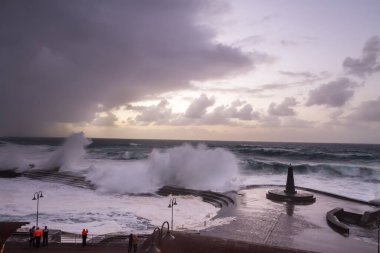 Büyük dalgaların fırtınası dalgakırana karşı kırılıyor ve Bajamar geziniyor gri fırtına bulutları ve arka planda bir deniz feneri ile.