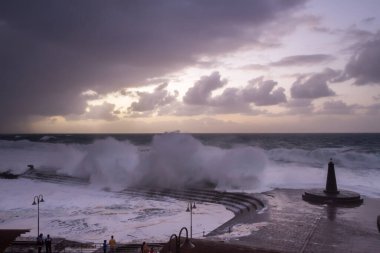 Büyük dalgaların fırtınası dalgakırana karşı kırılıyor ve Bajamar geziniyor gri fırtına bulutları ve arka planda bir deniz feneri ile.
