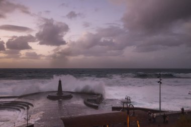 Büyük dalgaların fırtınası dalgakırana karşı kırılıyor ve Bajamar geziniyor gri fırtına bulutları ve arka planda bir deniz feneri ile.