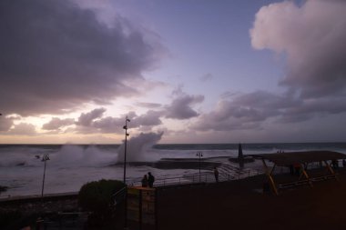 Büyük dalgaların fırtınası dalgakırana karşı kırılıyor ve Bajamar geziniyor gri fırtına bulutları ve arka planda bir deniz feneri ile.