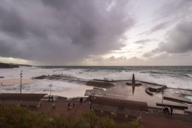 Büyük dalgaların fırtınası dalgakırana karşı kırılıyor ve Bajamar geziniyor gri fırtına bulutları ve arka planda bir deniz feneri ile.
