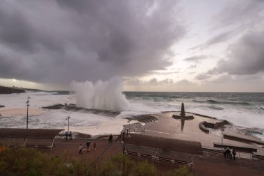 Büyük dalgaların fırtınası dalgakırana karşı kırılıyor ve Bajamar geziniyor gri fırtına bulutları ve arka planda bir deniz feneri ile.