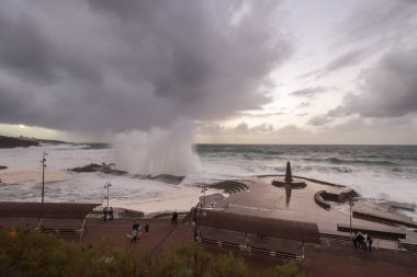 Büyük dalgaların fırtınası dalgakırana karşı kırılıyor ve Bajamar geziniyor gri fırtına bulutları ve arka planda bir deniz feneri ile.