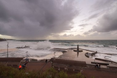 Büyük dalgaların fırtınası dalgakırana karşı kırılıyor ve Bajamar geziniyor gri fırtına bulutları ve arka planda bir deniz feneri ile.