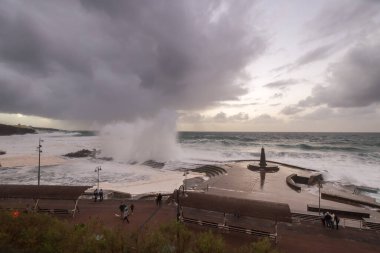 Büyük dalgaların fırtınası dalgakırana karşı kırılıyor ve Bajamar geziniyor gri fırtına bulutları ve arka planda bir deniz feneri ile.