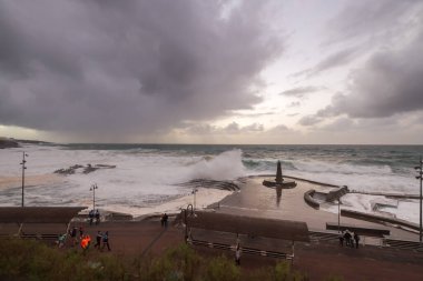 Büyük dalgaların fırtınası dalgakırana karşı kırılıyor ve Bajamar geziniyor gri fırtına bulutları ve arka planda bir deniz feneri ile.