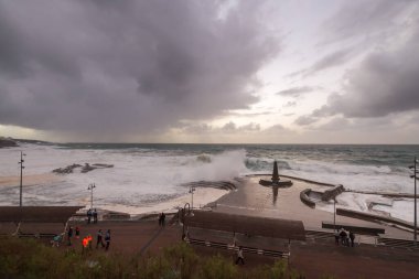 Büyük dalgaların fırtınası dalgakırana karşı kırılıyor ve Bajamar geziniyor gri fırtına bulutları ve arka planda bir deniz feneri ile.