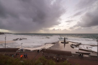 Büyük dalgaların fırtınası dalgakırana karşı kırılıyor ve Bajamar geziniyor gri fırtına bulutları ve arka planda bir deniz feneri ile.