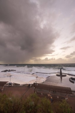 Büyük dalgaların fırtınası dalgakırana karşı kırılıyor ve Bajamar geziniyor gri fırtına bulutları ve arka planda bir deniz feneri ile.