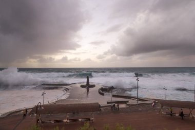 Büyük dalgaların fırtınası dalgakırana karşı kırılıyor ve Bajamar geziniyor gri fırtına bulutları ve arka planda bir deniz feneri ile.