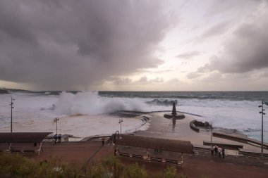Büyük dalgaların fırtınası dalgakırana karşı kırılıyor ve Bajamar geziniyor gri fırtına bulutları ve arka planda bir deniz feneri ile.