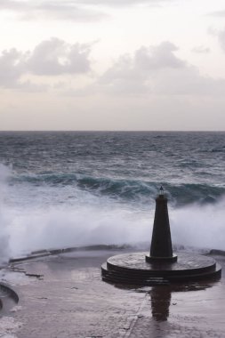 Büyük dalgaların fırtınası dalgakırana karşı kırılıyor ve Bajamar geziniyor gri fırtına bulutları ve arka planda bir deniz feneri ile.