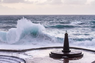 Büyük dalgaların fırtınası dalgakırana karşı kırılıyor ve Bajamar geziniyor gri fırtına bulutları ve arka planda bir deniz feneri ile.