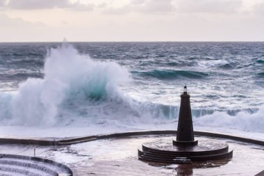 Büyük dalgaların fırtınası dalgakırana karşı kırılıyor ve Bajamar geziniyor gri fırtına bulutları ve arka planda bir deniz feneri ile.