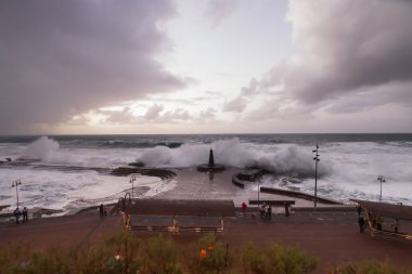 Büyük dalgaların fırtınası dalgakırana karşı kırılıyor ve Bajamar geziniyor gri fırtına bulutları ve arka planda bir deniz feneri ile.