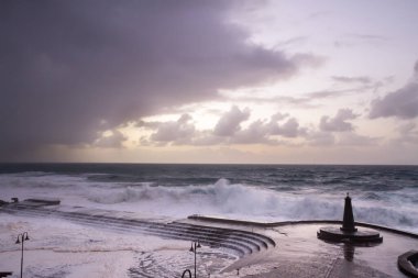 Büyük dalgaların fırtınası dalgakırana karşı kırılıyor ve Bajamar geziniyor gri fırtına bulutları ve arka planda bir deniz feneri ile.