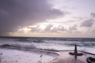 Büyük dalgaların fırtınası dalgakırana karşı kırılıyor ve Bajamar geziniyor gri fırtına bulutları ve arka planda bir deniz feneri ile.