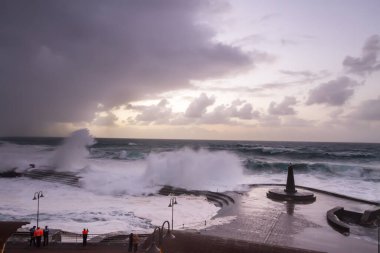 Büyük dalgaların fırtınası dalgakırana karşı kırılıyor ve Bajamar geziniyor gri fırtına bulutları ve arka planda bir deniz feneri ile.