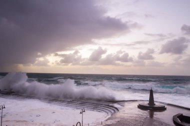 Büyük dalgaların fırtınası dalgakırana karşı kırılıyor ve Bajamar geziniyor gri fırtına bulutları ve arka planda bir deniz feneri ile.